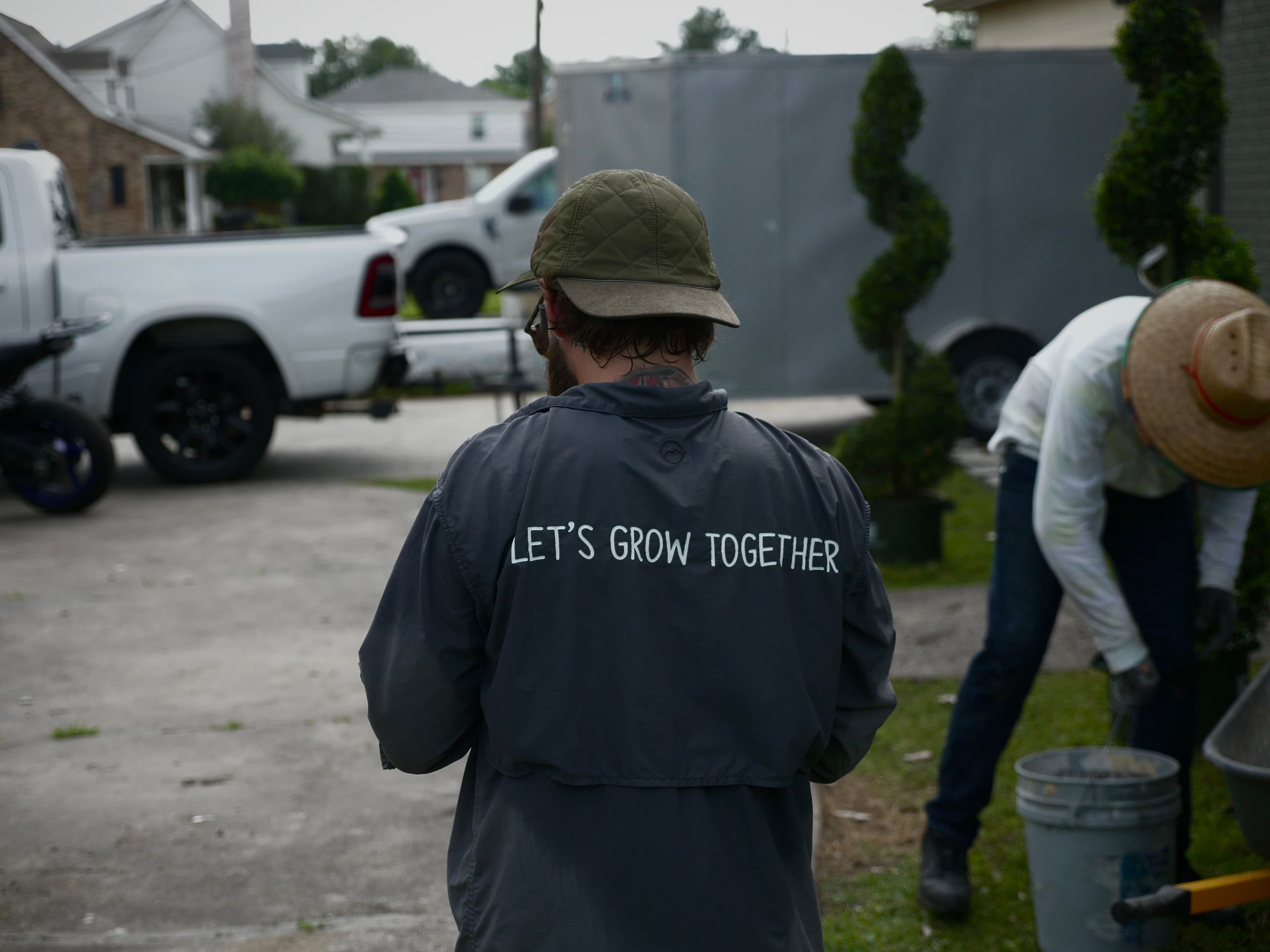 Person wearing a jacket with "LET'S GROW TOGETHER" text, working outdoors in a garden setting.