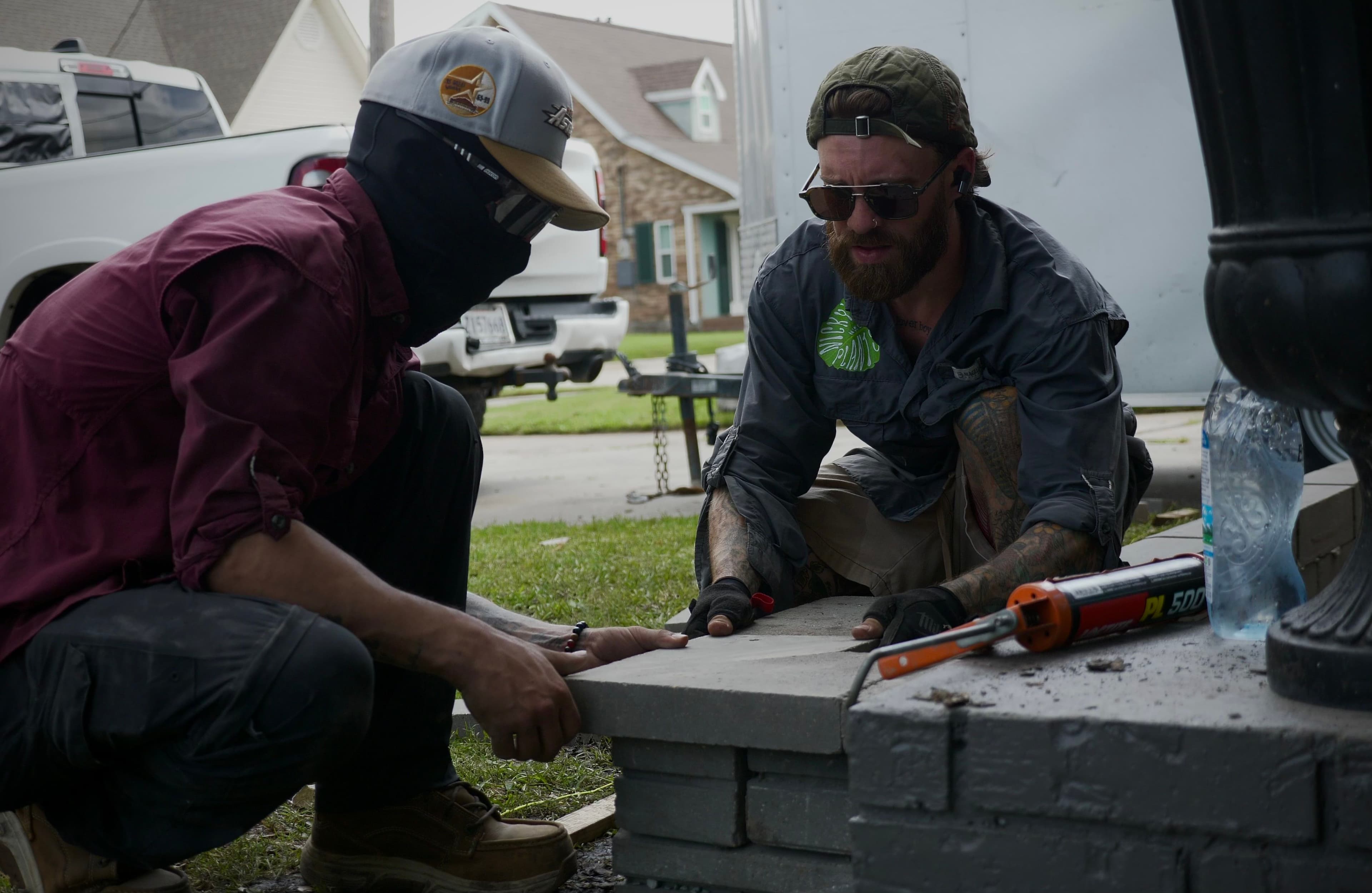 Two workers in protective gear laying bricks outdoors with tools nearby.